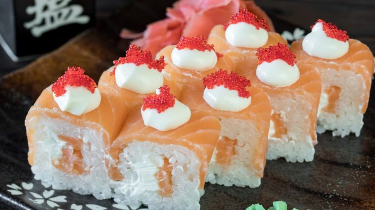 Close-up of a salmon sushi roll topped with cream cheese and red roe, garnished with wasabi.